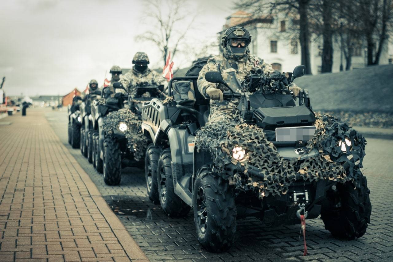 Četri ar karogiem rotāti transportlīdzekļi, uz kuriem sēž zemessargi pilnā kaujas ekipējumā, brauc pa Ventspils valstspilsētas ielu