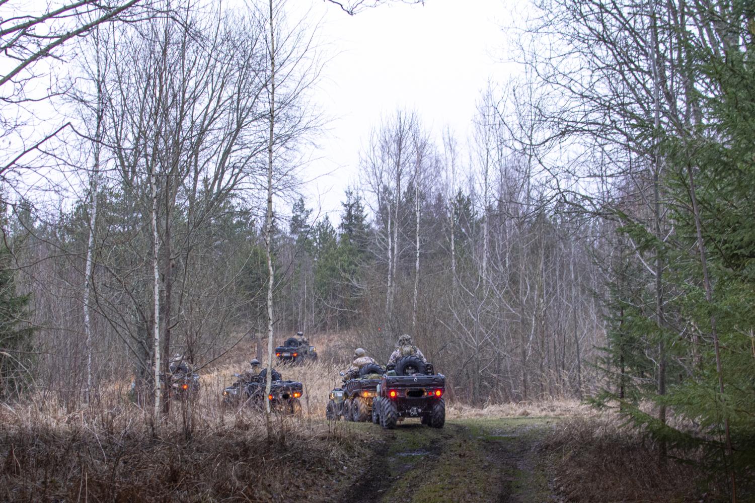 Pieci sešriteņu kvadricikli, uz kuriem sēž zemessargi pilnā kaujas ekipējumā, brauc mežainā apvidū